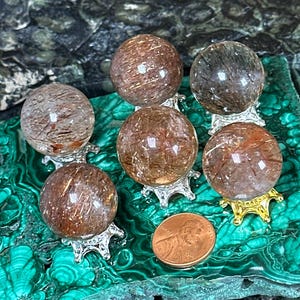 May include: Six crystal spheres, approximately 0.9 to 0.94 inches in diameter, rest on decorative stands. The spheres are a mix of brown and clear with internal inclusions. A penny is included for size reference. The spheres are displayed on a green and black surface.