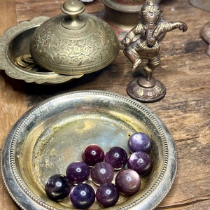 May include: A collection of polished purple fluorite spheres displayed in a tarnished silver dish. The spheres vary in shades of purple and are arranged on a wooden surface. Other items include a brass lidded bowl, a small brass statue, and a decorative bowl.