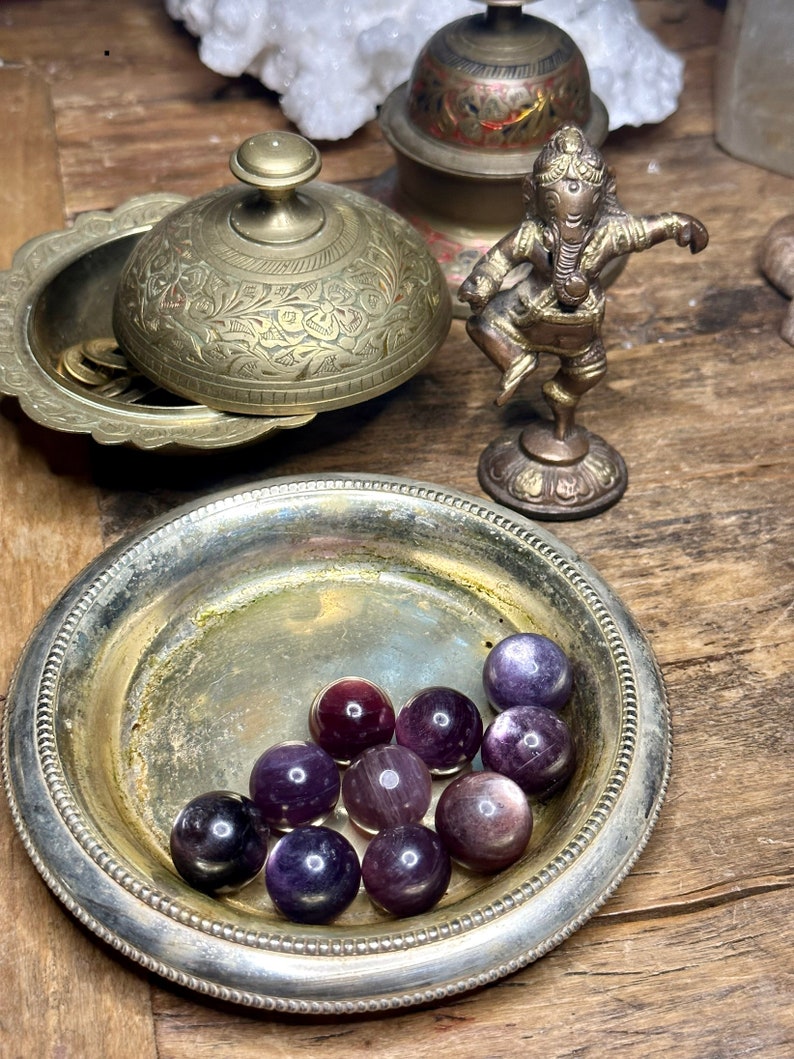May include: A collection of polished purple fluorite spheres displayed in a tarnished silver dish. The spheres vary in shades of purple and are arranged on a wooden surface. Other items include a brass lidded bowl, a small brass statue, and a decorative bowl.