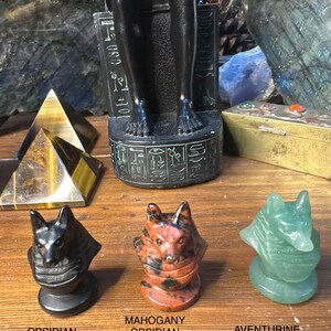 May include: Three carved stone fox head figurines in black obsidian, mahogany obsidian, and green aventurine. The figurines are on a wooden surface, with text labels below each one. A black statue and a gold pyramid are in the background.