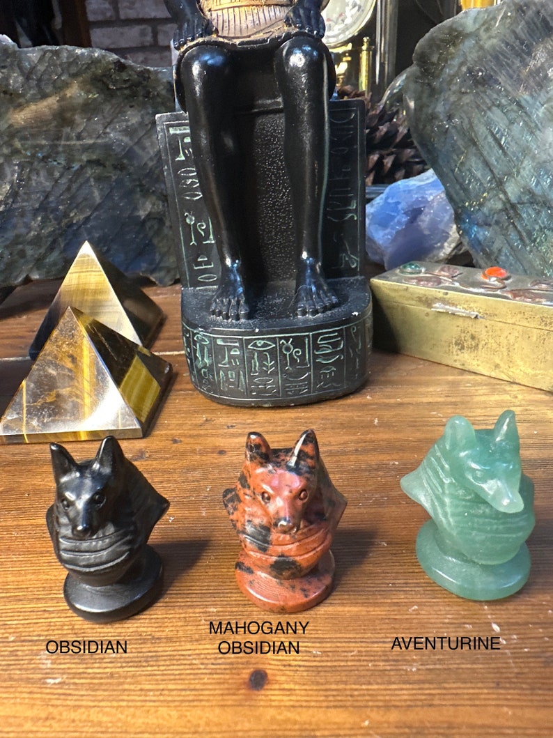 May include: Three carved stone fox head figurines in black obsidian, mahogany obsidian, and green aventurine. The figurines are on a wooden surface, with text labels below each one. A black statue and a gold pyramid are in the background.