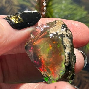 May include: A rough-cut opal gemstone with vibrant internal colors of red, green, and yellow. The stone has a dark, textured exterior and is held between fingers. The background is blurred.