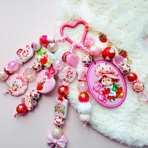 May include: A pink heart-shaped keyring featuring multiple strands of colourful beads, charms, and a large oval pendant depicting a character with a strawberry hat holding a pink animal. The accessory includes various strawberry-themed elements.