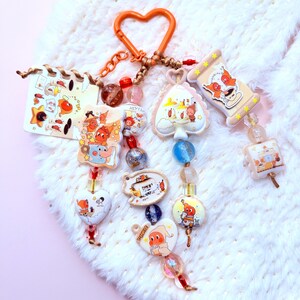 May include: A whimsical keychain featuring various charm shapes, including hearts, spades, and rectangles, adorned with cartoon characters and colorful beads. The keychain has an orange heart-shaped clasp and a mix of red, blue, and clear beads.