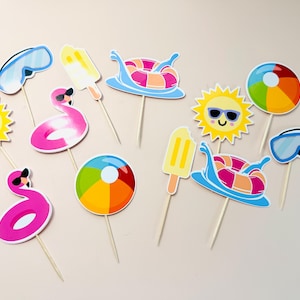 May include: A collection of summer-themed cupcake toppers featuring colorful illustrations of yellow suns with sunglasses, pink flamingo pool floats, blue swimming goggles, yellow popsicles, beach balls, and striped inner tubes on wooden sticks.