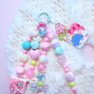 May include: A pastel-colored keychain with a variety of beads, including hearts, stars, and round shapes. The keychain features a light blue clasp, a small square with a cartoon illustration, and a bow. The color palette includes pink, blue, and green.