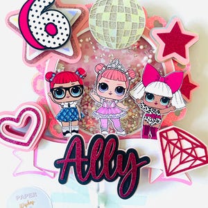 以下が含まれることがあります： 3体の漫画のドール、ディスコボール、数字の6が付いたピンクと白のケーキトッパー。トッパーには筆記体で「Ally」という名前とダイヤモンドの形が含まれています。背景は小さなピンク色のビーズで埋められています。