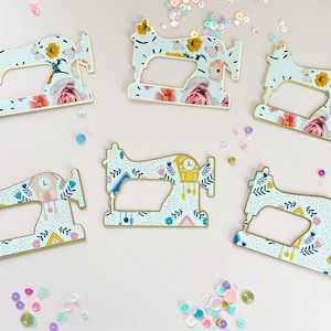 Puede incluir: Varios recortes decorativos de máquinas de coser con patrones florales y bordes dorados están dispersos sobre una superficie rosa claro. Los diseños incluyen rosas, mariposas y otros elementos botánicos. Lentejuelas de colores están esparcidas.