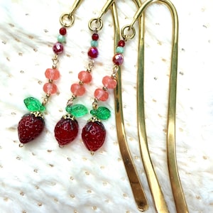 May include: Three gold-toned metal hook bookmarks, each featuring a dangling charm strand. The strands include small red and light green beads, two translucent peach-colored faceted beads, a green glass leaf, and a red strawberry-shaped bead.
