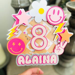 Könnte beinhalten: Ein rosa-weißer Tortendekor mit der Zahl 8, dem Namen Alaina und verschiedenen bunten Dekorationen, darunter Sterne, Blitze und Smiley-Gesichter.