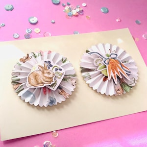Pode incluir: Duas decorações de rosetas de papel branco com enfeites temáticos de Páscoa. Uma apresenta um coelhinho marrom e detalhes florais, a outra tem cenouras e vegetação. As decorações estão em um cartão creme sobre um fundo rosa.