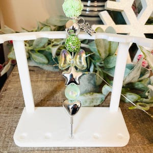 Puede incluir: Un tapón de botella decorativo con un diseño de corazón, estrella y lazo plateado. El tapón presenta una cuenta floral verde claro, una cuenta plateada y una bola de cuentas verdes. El tapón se exhibe en un soporte blanco.