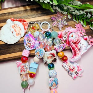 May include: A decorative keychain featuring a collection of colorful beads, a clear snowflake charm, and several holiday-themed acrylic pendants including Santa Claus, a snowman, a reindeer, a nutcracker, and winter houses in shades of pink, red, and blue.