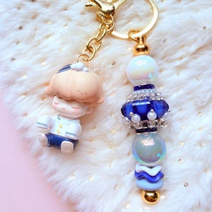May include: Two keychains with gold-colored hardware. One features a small figurine of a person in a white and blue outfit. The other has a blue and white beaded design with iridescent and gold accents. Both are on a fluffy white surface.