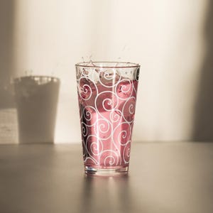 May include: A clear glass with a pink liquid and ice cubes, decorated with white swirling patterns. Water splashes from the top of the glass. The glass is on a reflective surface.