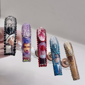 May include: A set of five long, embellished artificial nails. Each nail features a different design, including portraits, lace patterns, and gem accents. The nails are displayed on a clear stand against a white background.
