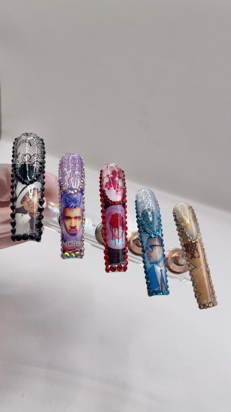 May include: A set of five long, embellished artificial nails. Each nail features a different design, including portraits, lace patterns, and gem accents. The nails are displayed on a clear stand against a white background.