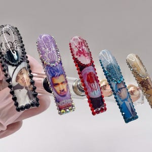 May include: Five long, embellished acrylic nails. Each nail features a different design, including portraits, lace, and gem accents. The nails are in various colors, including black, purple, red, blue, and tan, with a clear, glossy finish.