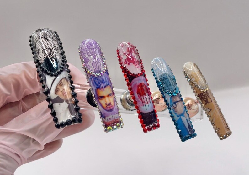 May include: Five long, embellished acrylic nails. Each nail features a different design, including portraits, lace, and gem accents. The nails are in various colors, including black, purple, red, blue, and tan, with a clear, glossy finish.