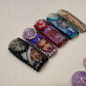 May include: A collection of decorative nail art designs. The designs include a black lace pattern, a photo, and various abstract patterns in shades of purple, red, and blue. Each nail is embellished with rhinestones.