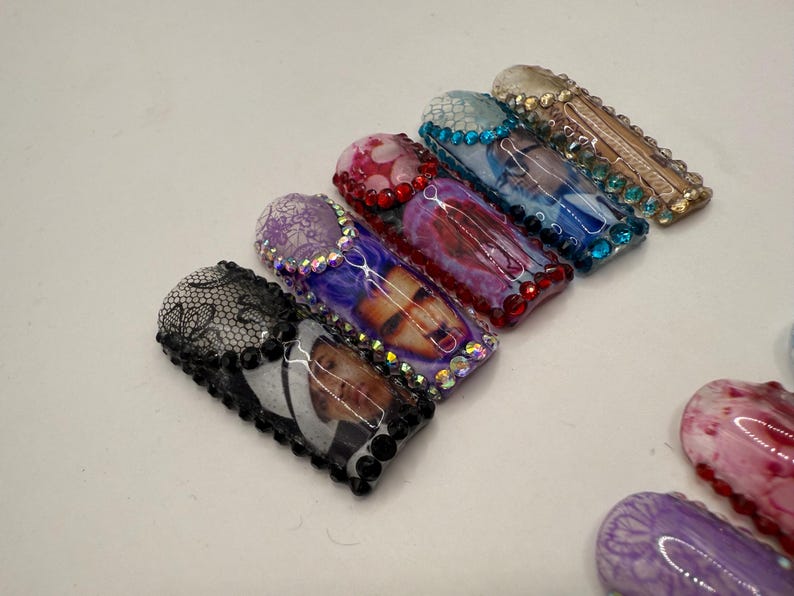 May include: A collection of decorative nail art designs. The designs include a black lace pattern, a photo, and various abstract patterns in shades of purple, red, and blue. Each nail is embellished with rhinestones.