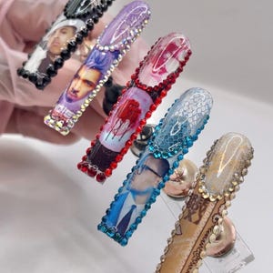May include: Five long, embellished acrylic nails. Each nail features a unique design, including portraits, lace, and floral patterns. The nails are adorned with rhinestones in various colors, creating a glamorous and artistic look.