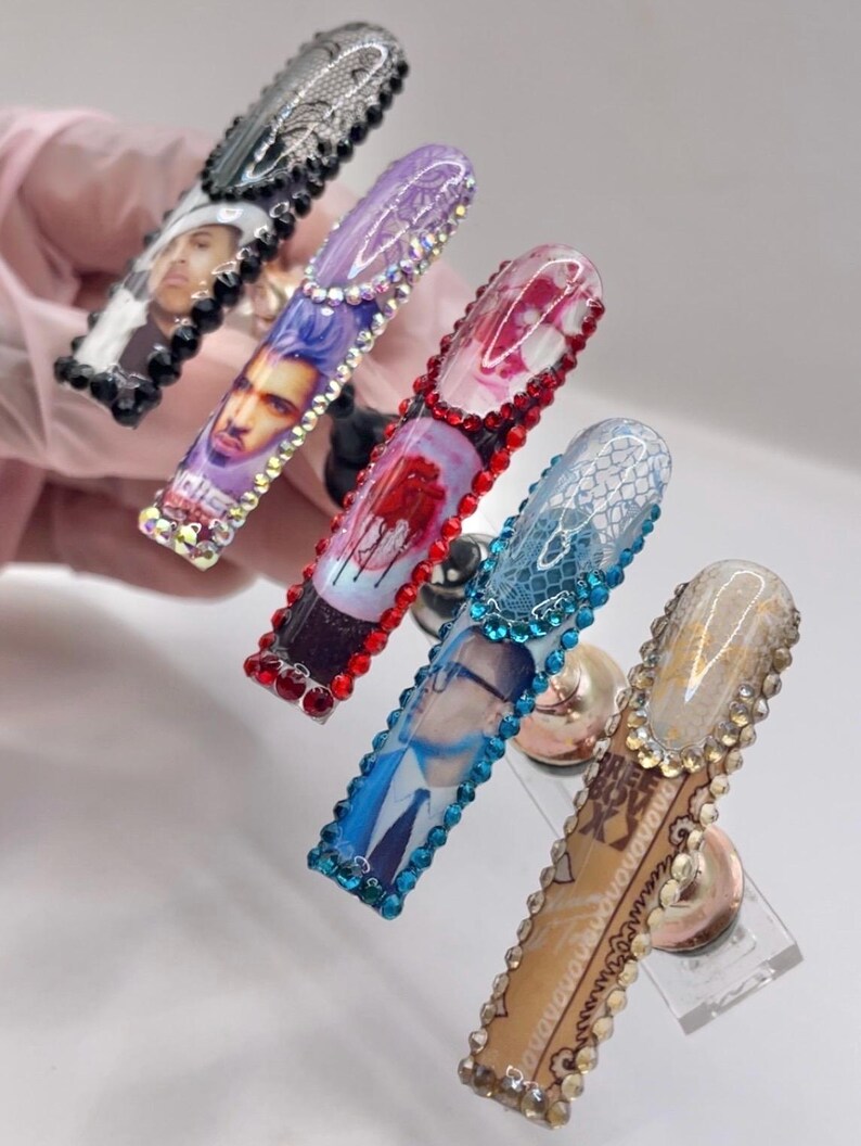 May include: Five long, embellished acrylic nails. Each nail features a unique design, including portraits, lace, and floral patterns. The nails are adorned with rhinestones in various colors, creating a glamorous and artistic look.