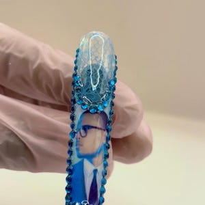 May include: A long, rectangular nail with a blue and white design. The top portion features a lace pattern, while the lower section displays a portrait of a person wearing glasses and a suit. The nail is adorned with blue rhinestones.