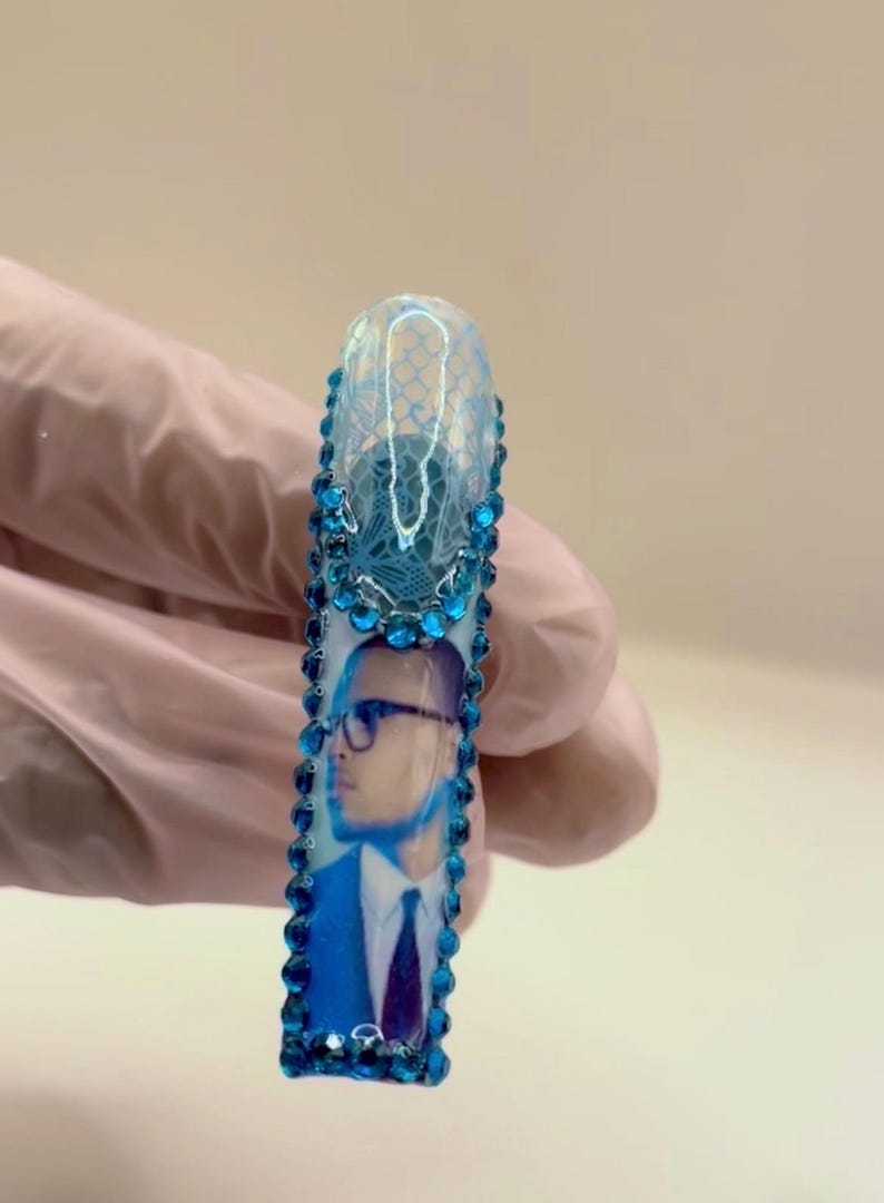 May include: A long, rectangular nail with a blue and white design. The top portion features a lace pattern, while the lower section displays a portrait of a person wearing glasses and a suit. The nail is adorned with blue rhinestones.