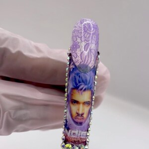 May include: A long, artificial nail with a design featuring a portrait of a man with blue hair and the word "Niche". The nail is decorated with rhinestones and a purple lace pattern.