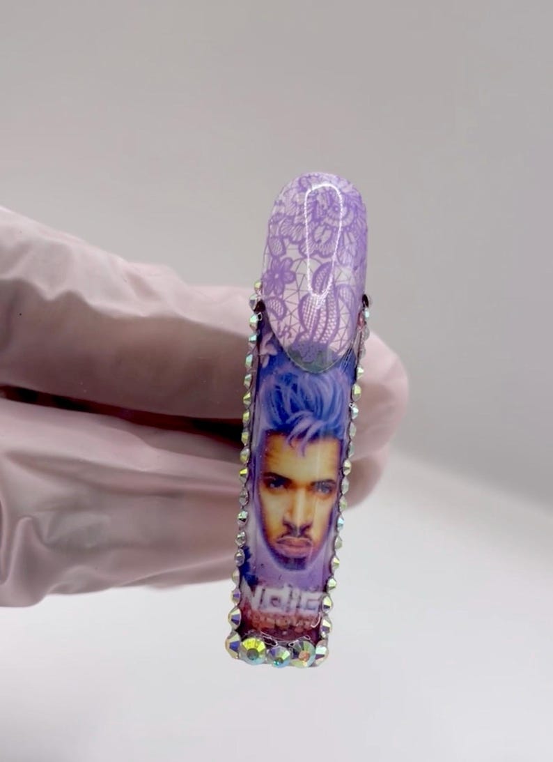May include: A long, artificial nail with a design featuring a portrait of a man with blue hair and the word "Niche". The nail is decorated with rhinestones and a purple lace pattern.