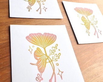 Puede incluir: Tres tarjetas cuadradas con un diseño botánico. Cada tarjeta presenta una ilustración estilizada de una hoja de ginkgo y otros elementos vegetales en tonos amarillos y rosas. Las tarjetas están dispuestas sobre una superficie de madera.