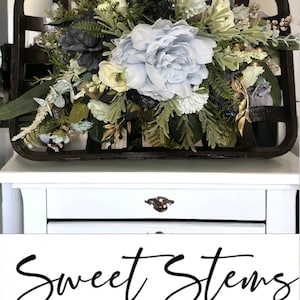 May include: A rustic wooden basket filled with a floral arrangement featuring blue, white, and green flowers. The arrangement includes peonies, hydrangeas, and greenery. The basket is sitting on a white table with a drawer. The text "Sweet Stems Wreath Company" is visible below the image.