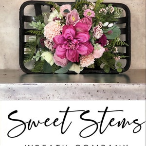 May include: A black basket filled with a pink, white, and green floral arrangement. The flowers are arranged in a bouquet style and include pink roses, white hydrangeas, and green ferns. The basket is sitting on a gray surface. The text "Sweet Stems Wreath Company" is displayed below the image.