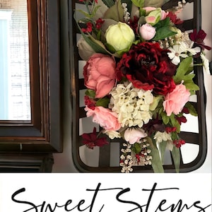 May include: A rustic wooden basket filled with a bouquet of artificial flowers. The flowers are in shades of pink, red, and white. The basket is hanging on a wall and has the text "Sweet Stems Wreath Company" below it.