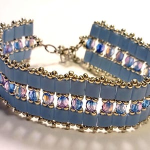 Peut inclure: Un bracelet bleu clair composé de perles carrées disposées en trois rangées. Le bracelet est orné de perles irisées bleues, violettes et roses. Les bords sont bordés de petites perles dorées et il possède un fermoir argenté.