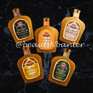 May include: Five gold-colored glass bottles with different Crown Royal flavors: Black, Peach, Vanilla, Regal Apple, and a fifth flavor that is not visible in the image. Each bottle has a gold crown and a label with the flavor name.