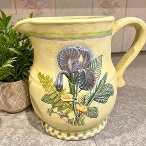 May include: A pale yellow ceramic pitcher featuring a raised, three-dimensional relief of a purple iris flower, green fern leaves, and small yellow blossoms. The pitcher has a crackled glaze finish, a curved handle, and a checkered base pattern.