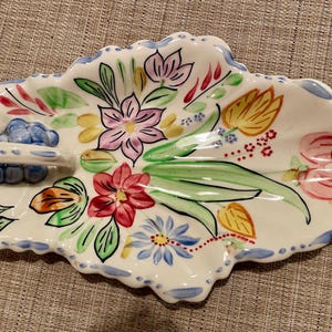May include: A leaf-shaped ceramic serving dish with a handle, hand-painted with colorful flowers and green leaves. The rim is scalloped and trimmed with blue paint. The dish measures approximately 12 inches long.