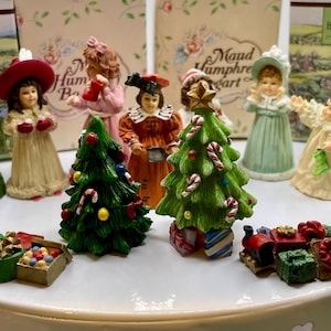 May include: A collection of Christmas figurines, including children in festive attire, Christmas trees, and a small train. The figurines are painted in bright colors and are arranged on a white surface. The background includes decorative cards.