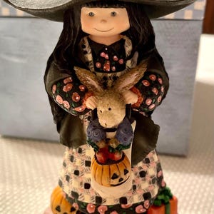 May include: A ceramic Halloween figurine of a witch holding a bunny and a pumpkin-shaped basket. The witch wears a black hat, a black coat with floral accents, and a patterned dress. The figurine is on a brown base with small pumpkins and a gift box.