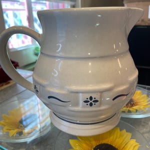 May include: A white ceramic pitcher with a blue and white floral design. The pitcher has a wide mouth and a handle. It is sitting on a table with a sunflower in the foreground.