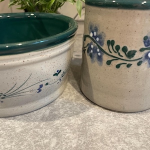 May include: A speckled beige ceramic bowl and a matching tall mug, both featuring a dark green rim and a hand-painted floral vine pattern in blue and green. The ceramic pieces have a smooth, speckled finish and a consistent design aesthetic.
