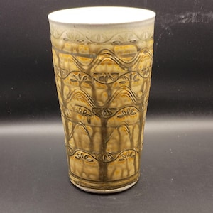 May include: A tall ceramic tumbler featuring a tapered cylindrical shape with a white interior rim. The exterior surface displays a repeating embossed pattern of dark brown wavy lines and floral motifs over a mottled tan and yellow glaze.
