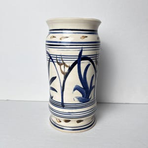 May include: A ceramic vase with a cream-colored base and a flared top. The vase is decorated with blue and brown brushstrokes and horizontal blue stripes. The vase has a footed base and is approximately 8 inches tall.