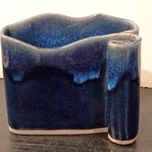 May include: A ceramic vessel with a deep blue glaze featuring a glossy finish and lighter blue drips along the rim. The object has an irregular, wavy top edge and a distinct cylindrical extension on the right side, with a visible light-colored base.