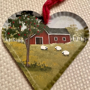 May include: Heart-shaped ornament with a painted pastoral scene. A red barn and apple tree are depicted in a field with three sheep. The ornament has a red yarn loop for hanging.