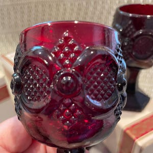 May include: Close-up of two ruby red wine glasses. The glasses have a detailed, embossed pattern on the bowl and a short stem. The design includes diamond and oval shapes. The glasses are likely vintage or antique.