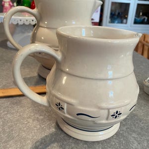 May include: Two cream-colored ceramic pitchers with blue floral designs. The larger pitcher has a wider base and a spout. The smaller pitcher has a narrower base and a more rounded shape.