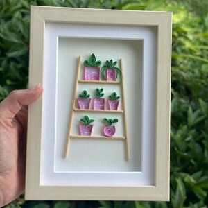 Peut inclure: Une œuvre d'art en quilling encadrée représentant une échelle miniature avec des pots de fleurs roses. Les pots contiennent des plantes en papier vert. Le cadre est en bois clair et le fond est de couleur blanc cassé.